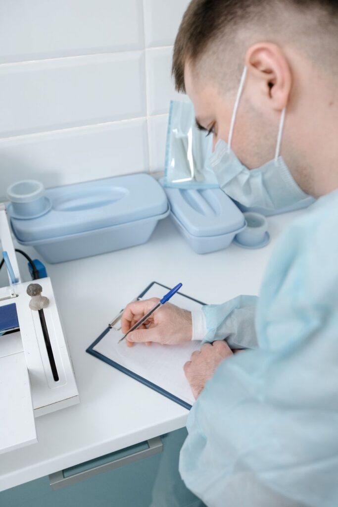 A medical professional wearing a mask writes notes on a clipboard in a hospital setting.